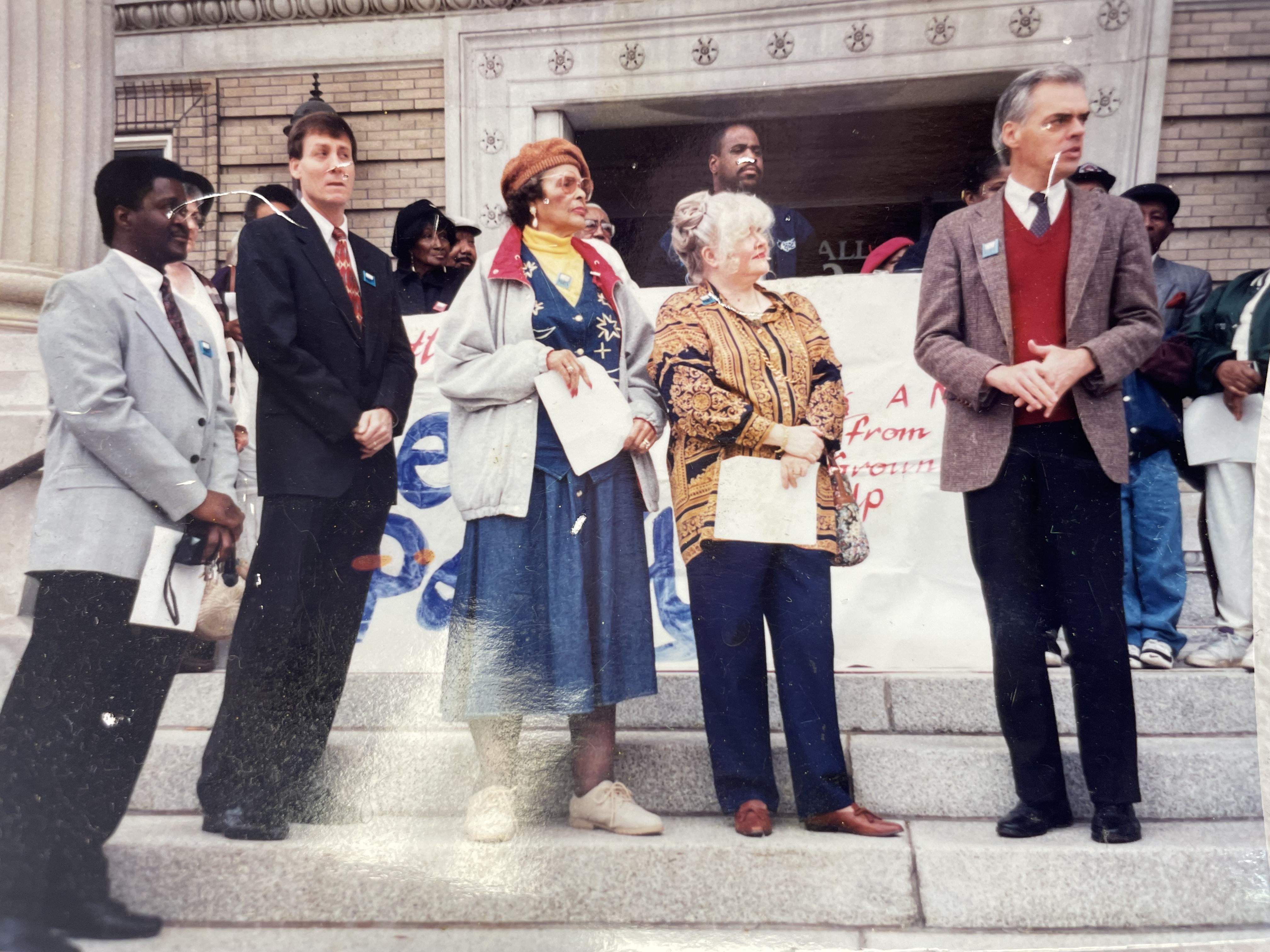 Photograph on the wall at the Arkansas Community Organizations (ACO) office in Little Rock, Arkansas. It’s of a press conference in the late 90s on the steps of Little Rock City Hall featuring New Party public officals. From left to right: Michael Daugherty, a Little Rock New Party member who was elected to the Little Rock School Board in 1995 and was re-elected for several terms of office until he was defeated in 2010; Paul Kelly, a Little Rock New Party member who was elected to the Little Rock City Board (City Council) in the Fall of 1996 and served a single four-year term; Johnnie Pugh, a longtime ACORN member and Co-Chairperson of the Little Rock New Party; Kathy Wells, a Little Rock New Party member who was very active in the city’s grassroots politics; and Jim Lynch, a longtime supporter and volunteer for ACORN and Co-Chairpeson of the Little Rock New Party. Each indivual is wearing a blue “New Party” button on their coats.