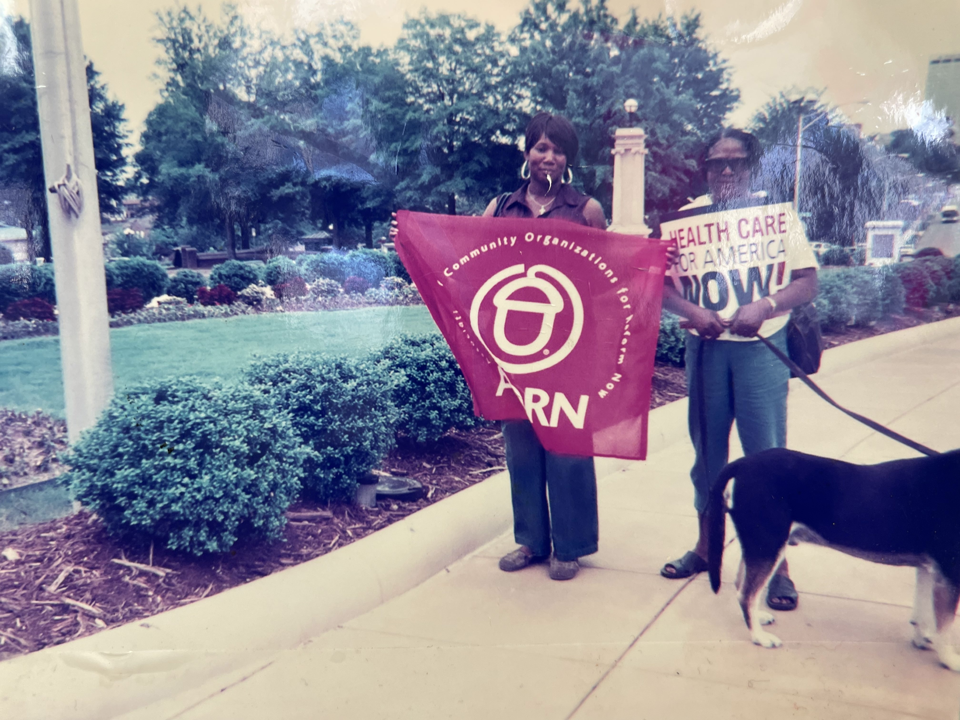 Photograph from the Arksansas Community Organizations (ACO) office in Little Rock, Arkansas, featuring two women holding up an ACORN flag and a poster that reads, “Health Care For America Now!”